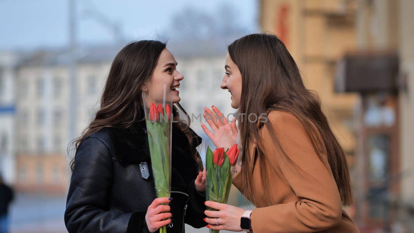 two happy girls, students, friends with flowers in their hands, laughing on a city street