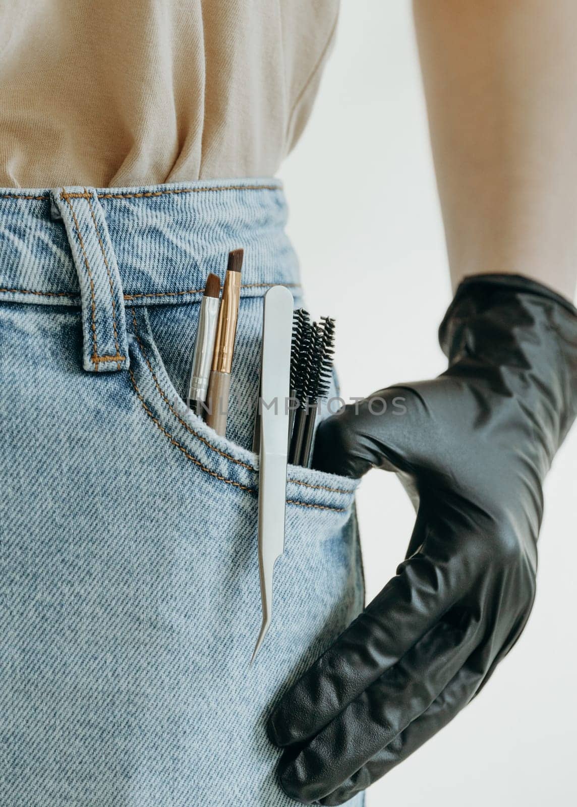 A set of brushes, tweezers and brushes for eyelash extensions stick out from the left pocket of blue jeans with a girl s hand in black gloves, close-up side view.