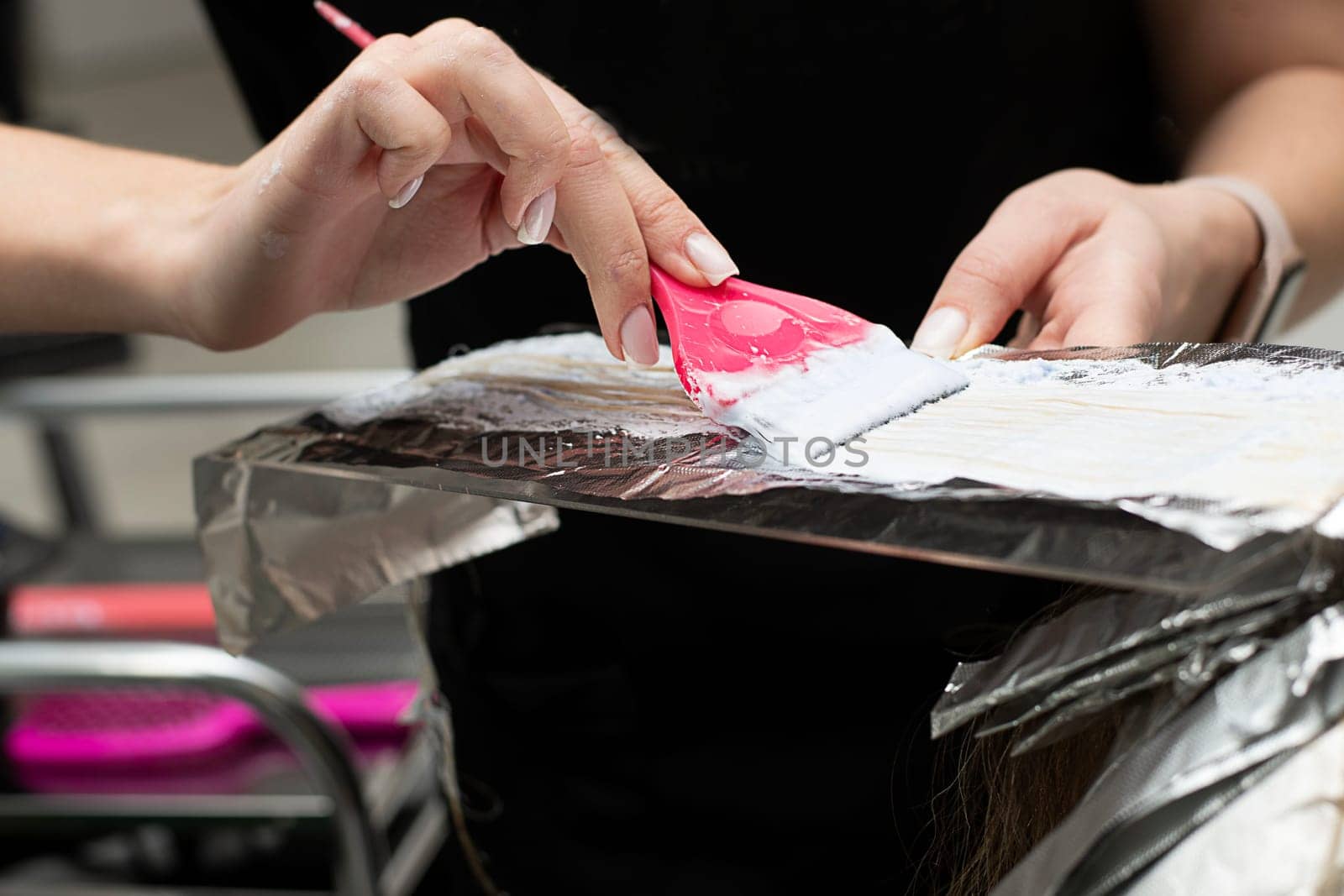 Hair coloring in a beauty salon. A master hairdresser-colorist dyes a client's brown hair blond. Apply lightening powder to hair onto foil using a pink brush. Close-up. Business concept.