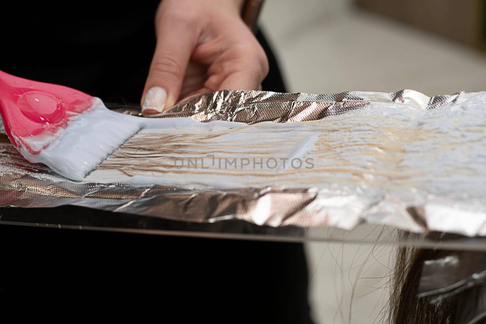 Hair coloring in a beauty salon. A master hairdresser-colorist dyes a client's brown hair blond. Apply lightening powder to hair onto foil using a pink brush. Close-up. Business concept.