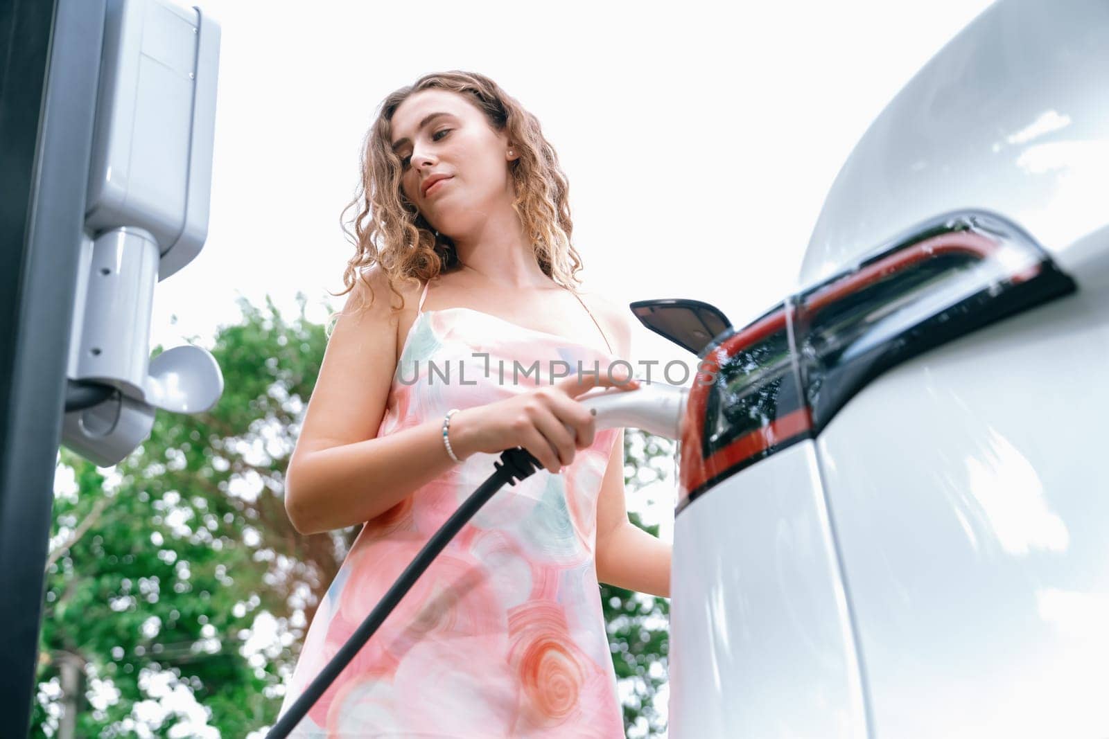 Eco-friendly conscious woman recharging modern electric vehicle from EV charging station. EV car technology utilized as alternative transportation for future sustainability. Synchronos