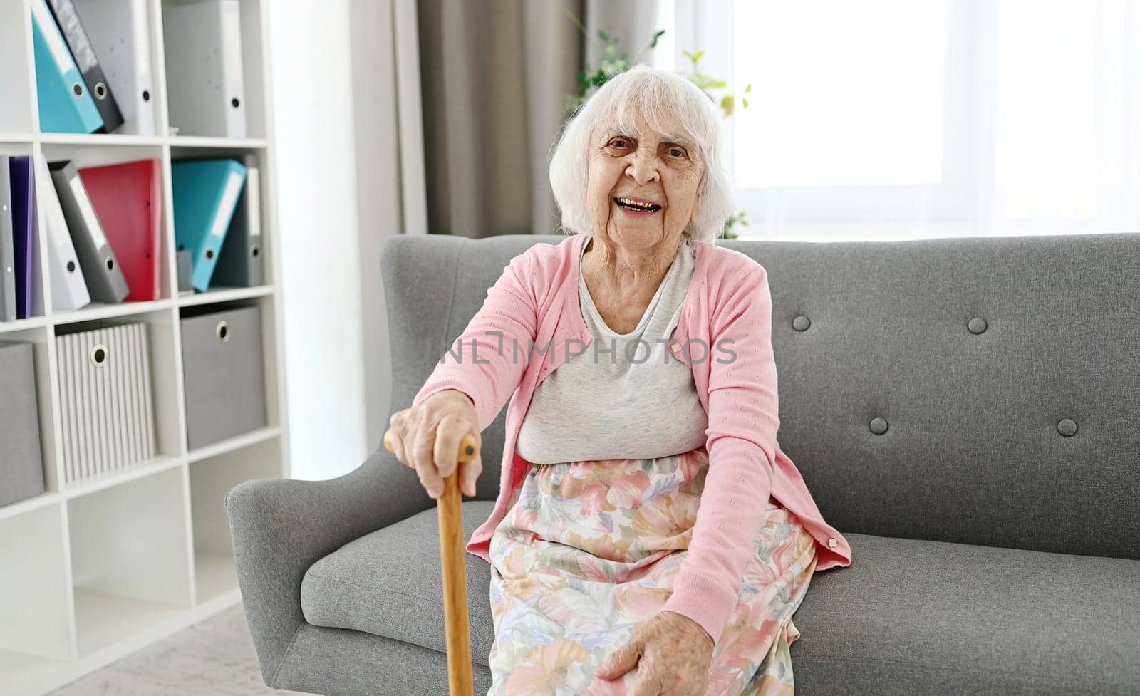Elderly Woman Sitting On A Living Room Sofa With Cane Laughing And Looking Into The Camera