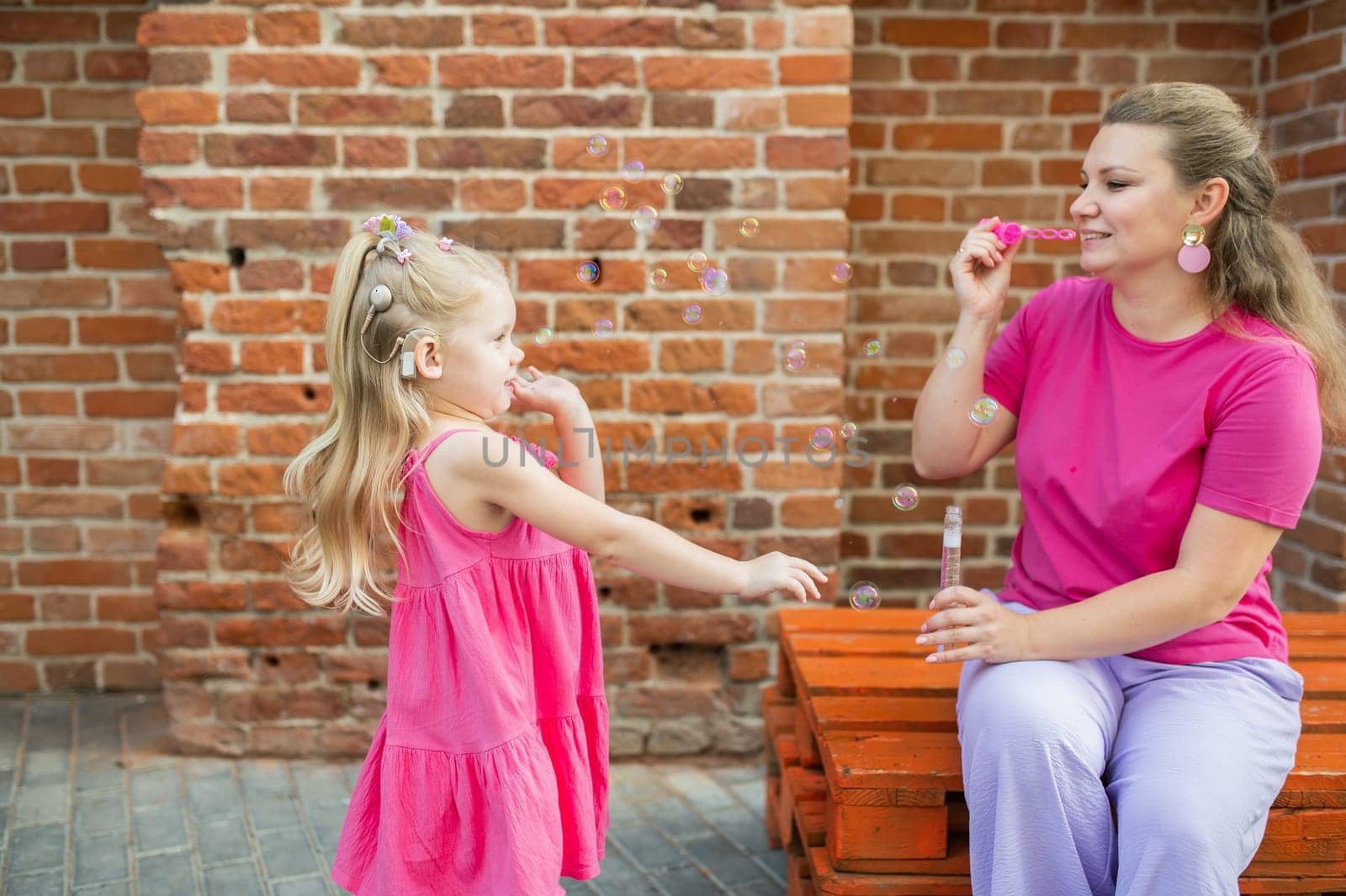 Deaf child with cochlear implant for hearing audio and aid for impairment having fun and laughs with mother outdoor in summer. Sound fitting device to help with communication listening and interaction.