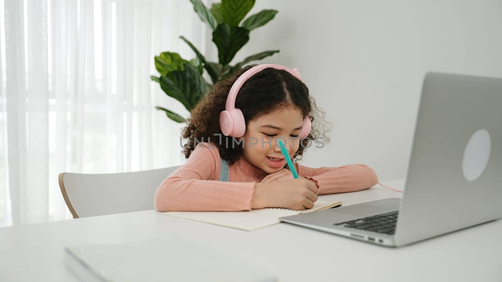 Cute american girl wearing headphone and doing homework in self study. Happy attractive schoolgirl writing at notebook while searching about programing system and writing engineering prompt. Pedagogy.