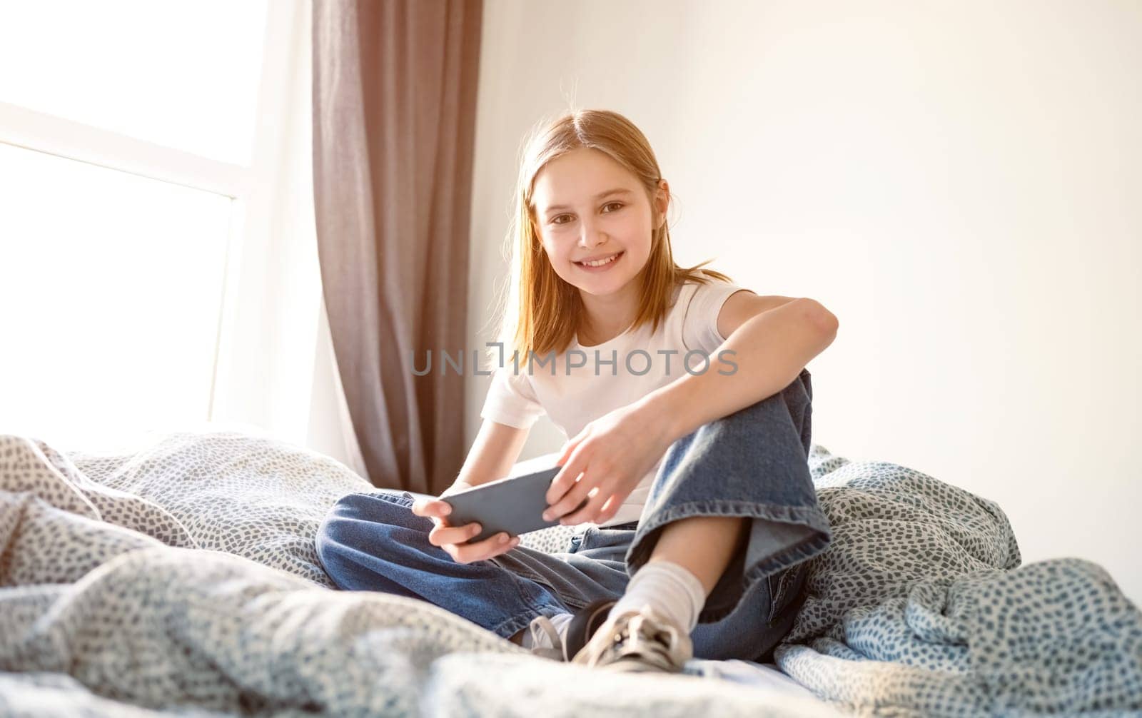 Cute Little Playing Game On A Smartphone Sitting On A Bed In Kid'S Room