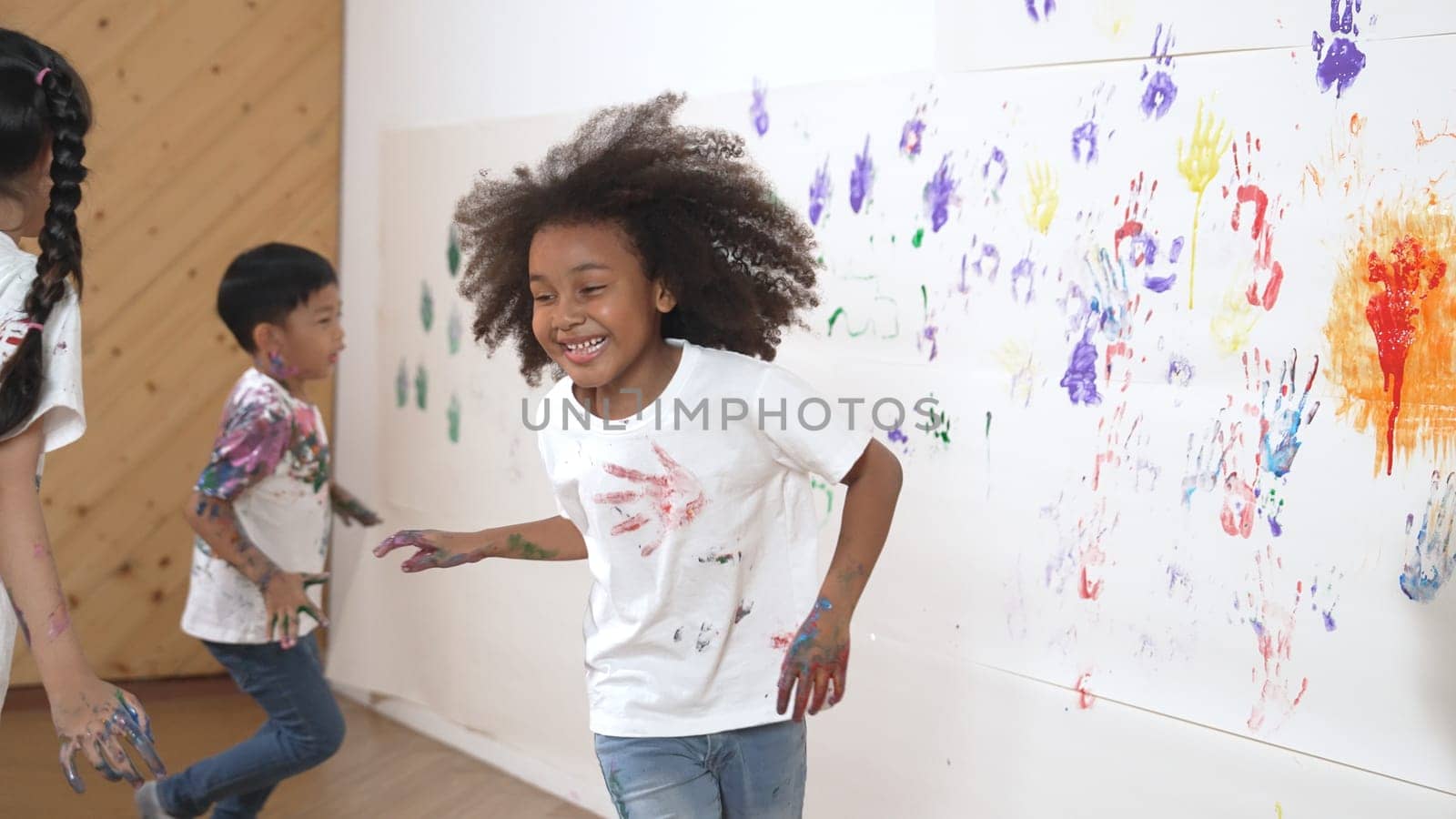 Young asian boy running to stamp colored hand print at cute african girl shirt. Student playing in art lesson with white background stained with colorful color. Creativity activity concept. Erudition.