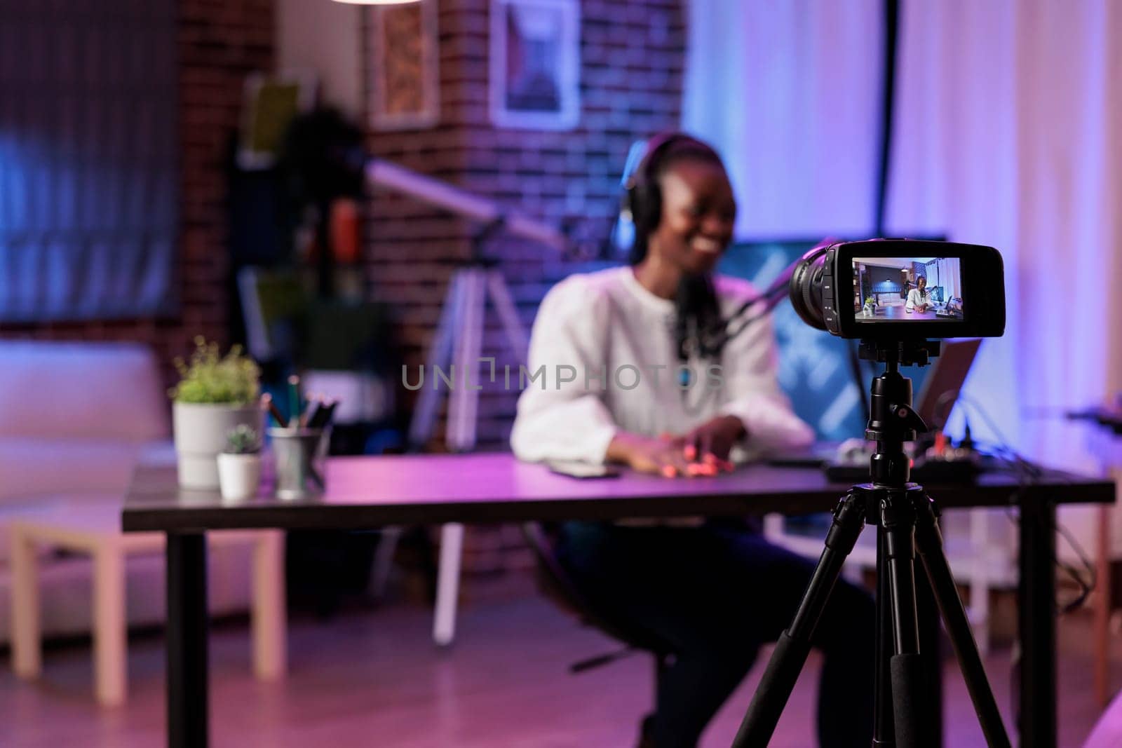 Focus shot on modern camera recording vlogger in blurry background talking about family and relationships subjects. Woman producing online show using high quality equipment in rgb lights studio
