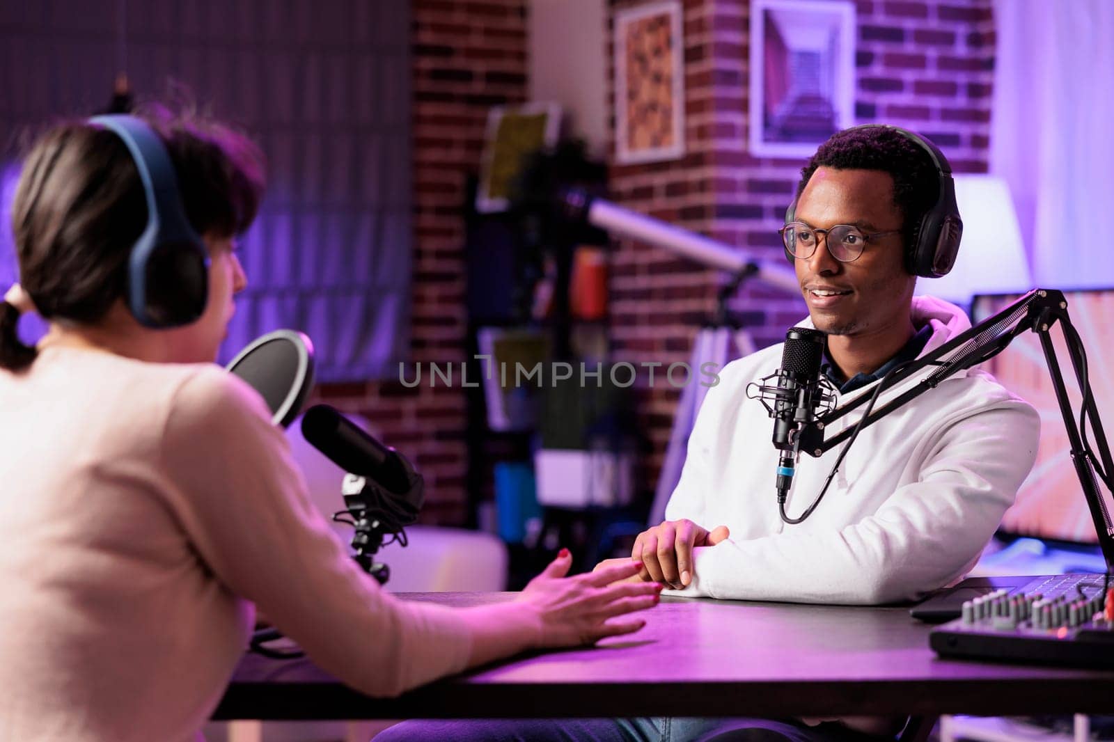 Man and woman broadcasting discussion on online podcast episode using professional equipment . Vlogger show host and guest talking in neon lights ornate living room personal studio broadcast