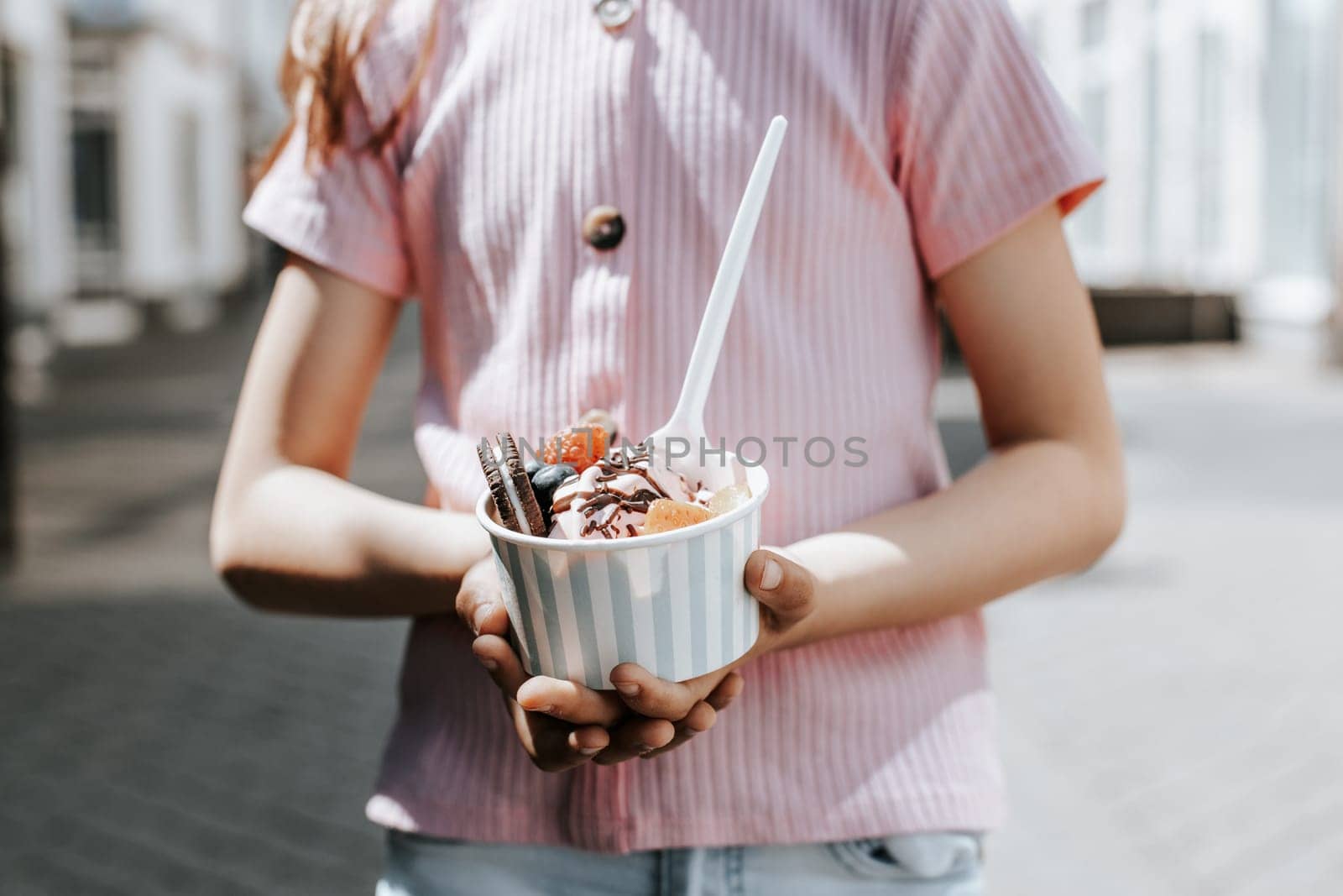 Portrait of one beautiful Caucasian unrecognizable girl holding a paper cup with ice cream, berries and cookies in her hands while standing on a city street on a clear sunny day,