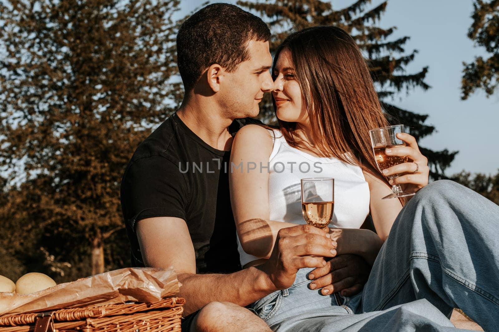 One beautiful caucasian young couple looks at each other with a tender look, holding glasses of champagne, sit on a bedspread with a wicker basket and fruits in the park on a summer sunny day, close-up side view.