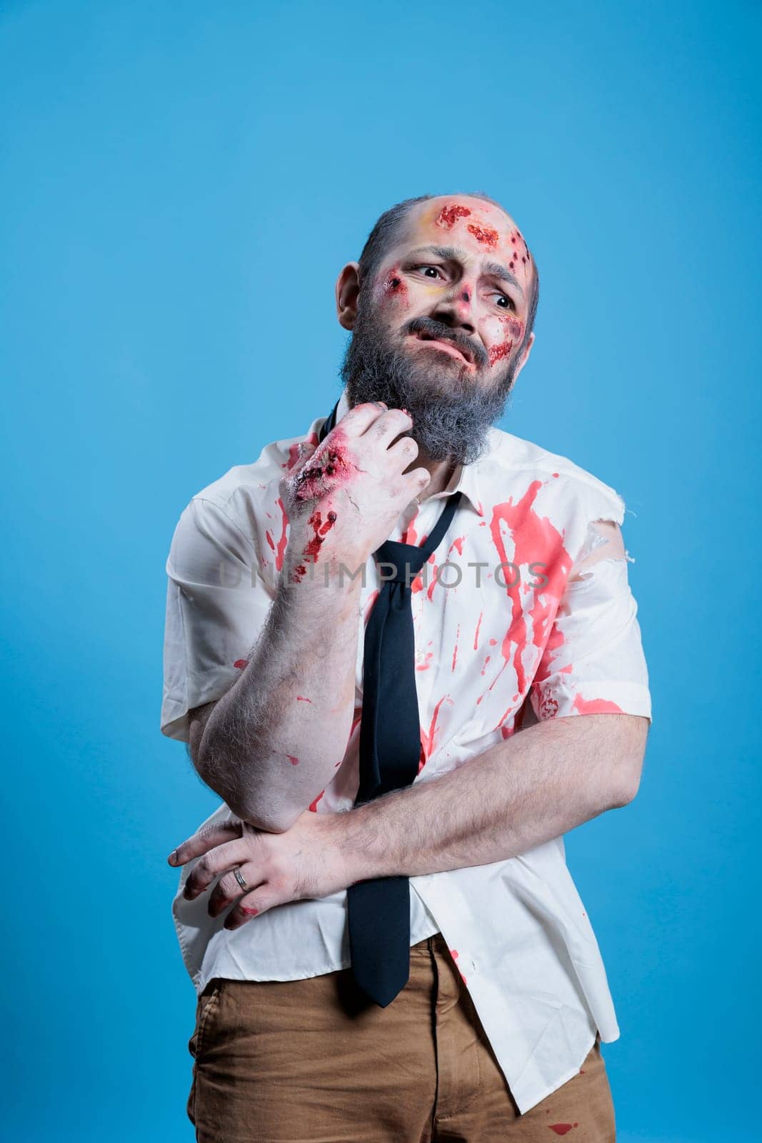 Portrait of man dressed as zombie wincing from pain for Halloween event, wearing horror makeup. Person costumed as infected diseased creature covered in blood and wounds, studio background