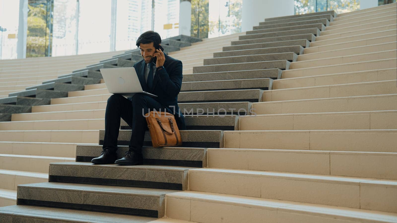 Skilled investor working or planing strategy by using laptop at stair. Professional business man wearing suit while working and typing data analysis by using laptop and talking to phone. Exultant.