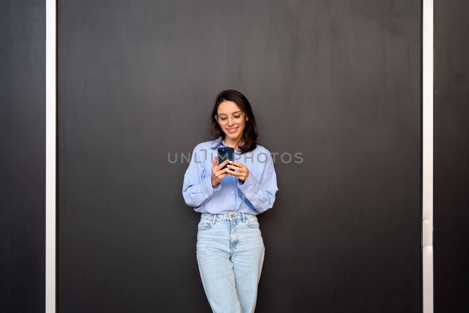 Satisfied hipster girl wear glasses, types text message on mobile phone, enjoys online communication, types feedback, wears denim shirt, isolated on black studio background. Technology concept