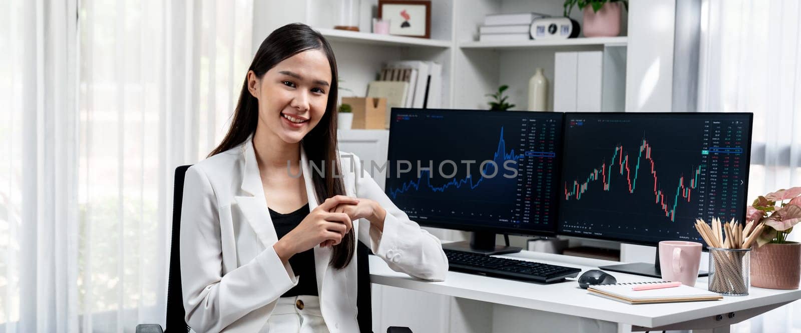 Profile of young Asian businesswoman smiling on happy face with white suit sitting chair against dynamic stock exchange market investing graph on pc showing screen on desk at modern office. Stratagem.