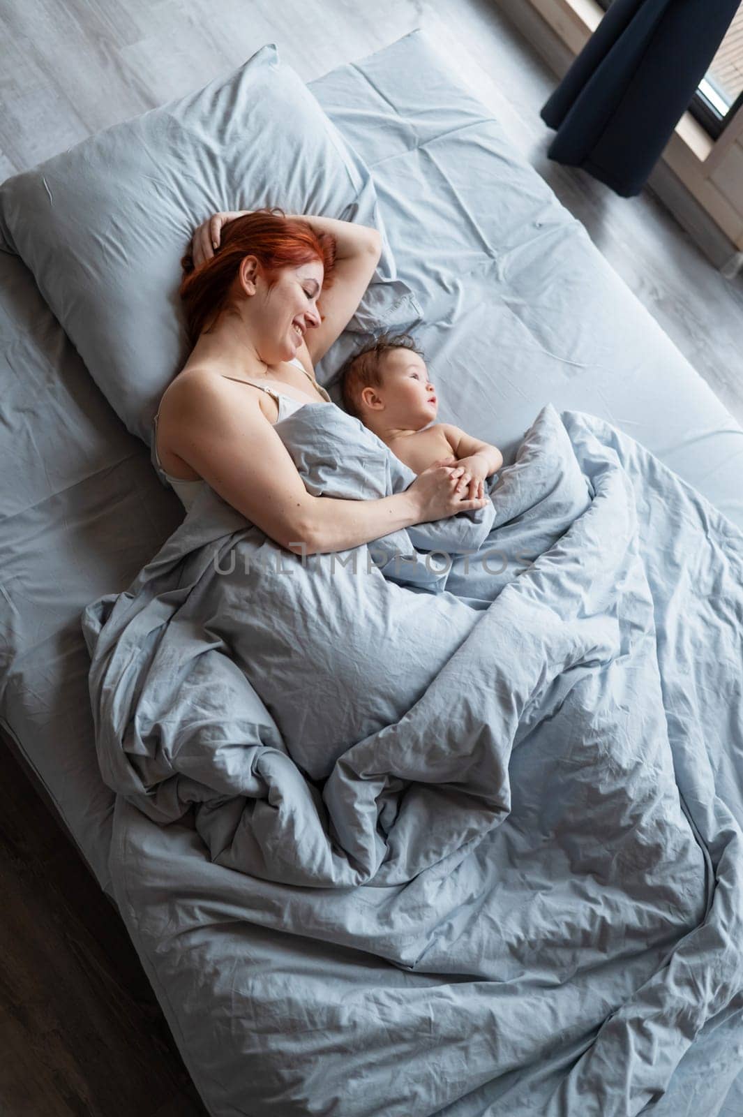 Top view of a red-haired Caucasian woman lying in bed with her baby son. Vertical photo. by mrwed54