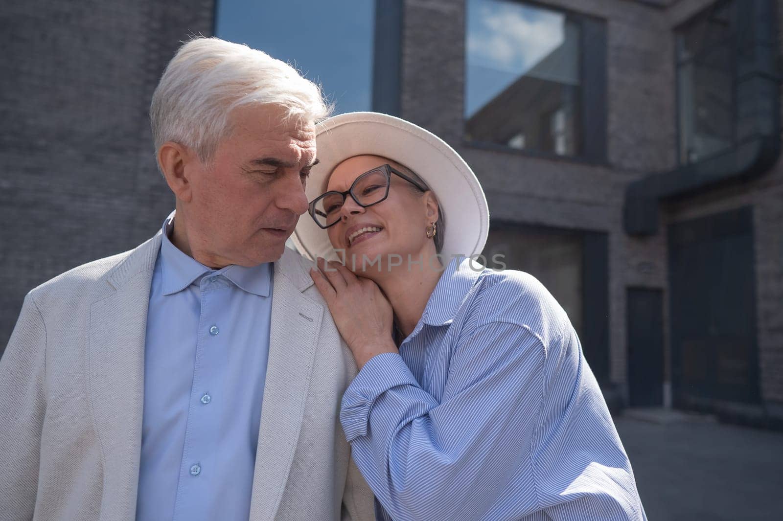 A woman in a hat and glasses hugs an elderly man in a white jacket from behind. Romantic relationships of mature people. by mrwed54