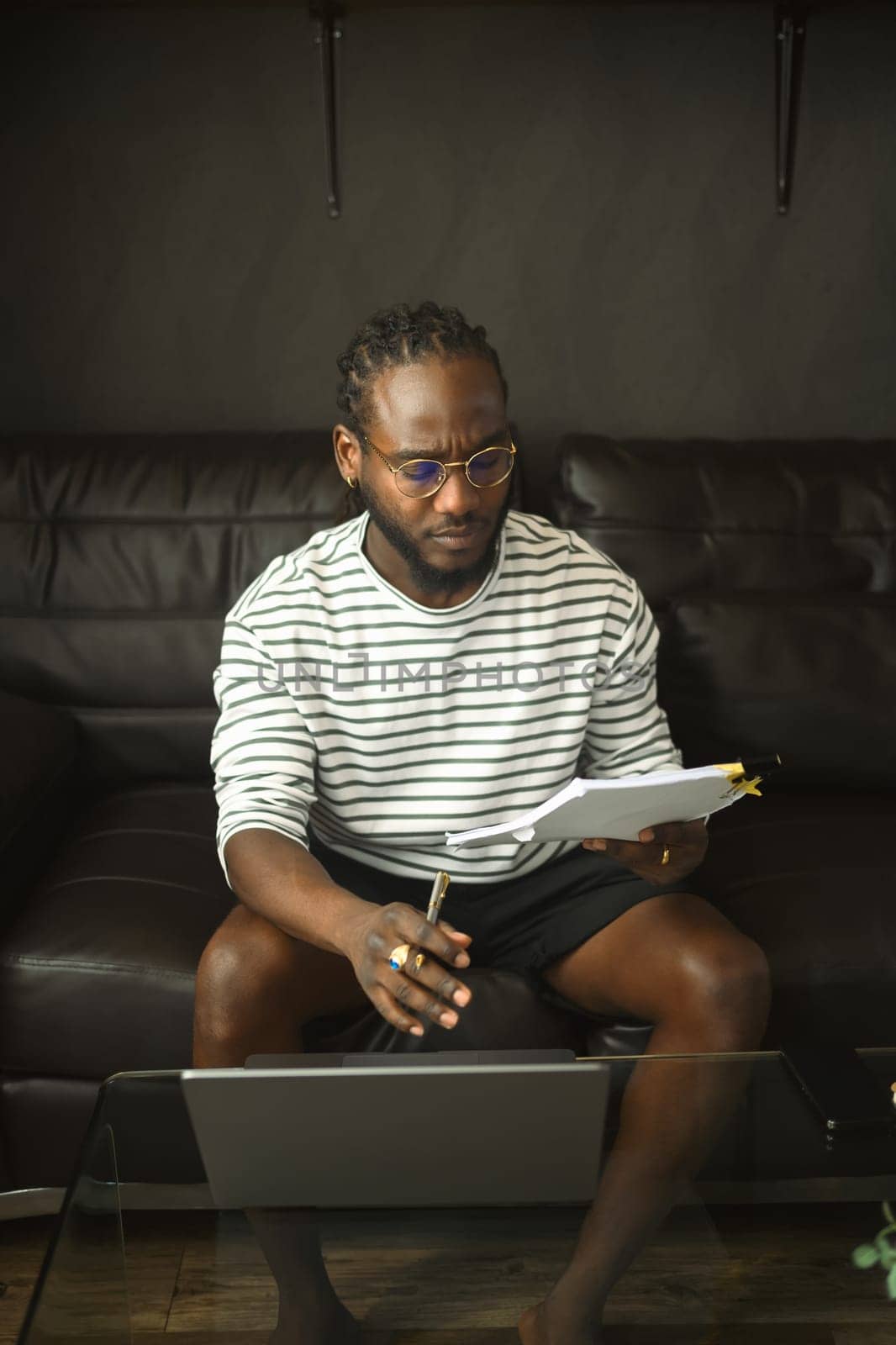 Focused African man analyzing documents while working from home on laptop.