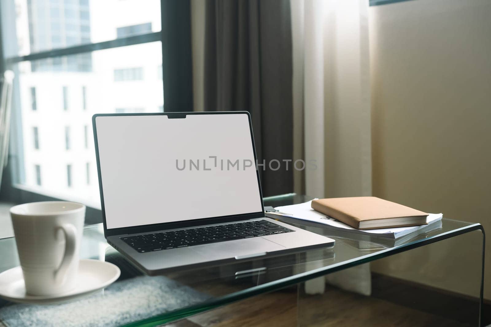 Laptop with white empty screen, notebook and coffee cup on glass table in living room.