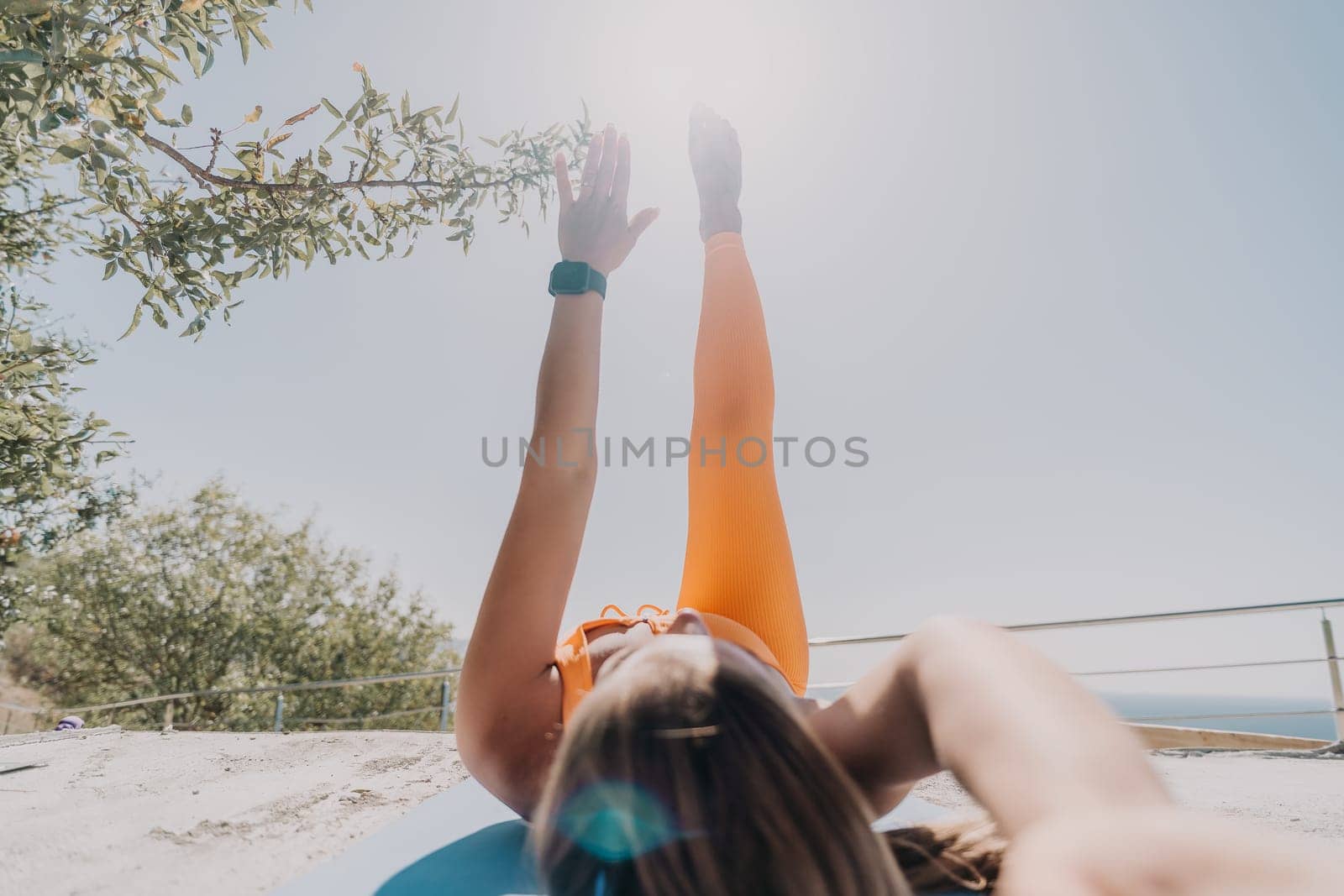 Fitness woman sea. Happy middle aged woman in orange sportswear exercises morning outdoors on yoga mat with laptop in park over ocean beach. Female fitness pilates yoga routine. Healthy lifestyle. by panophotograph