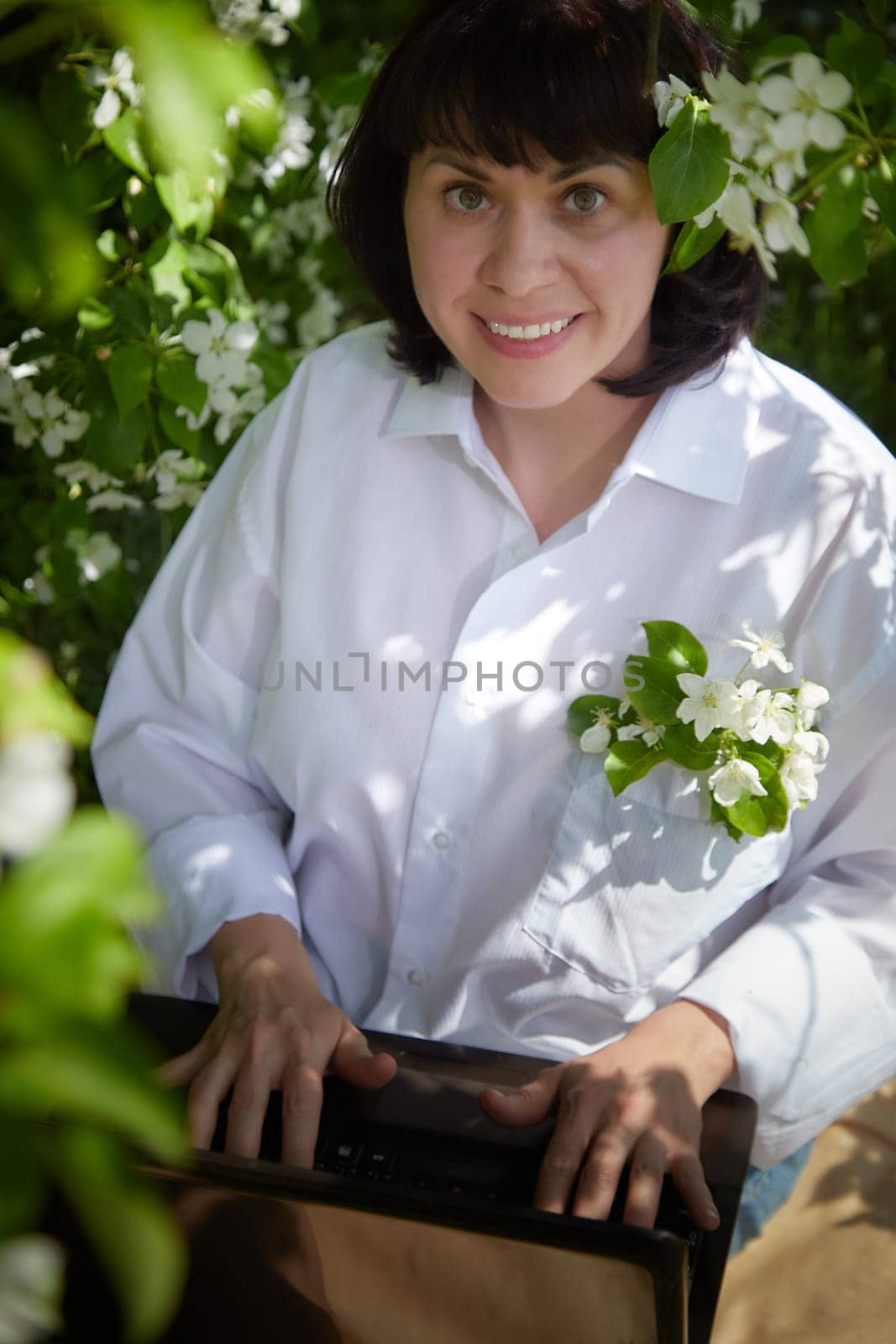 Girl Working on Laptop in Blossoming Garden. Middle aged woman using computer near white apple blossoms. Female freelancer, blogger working outdoors