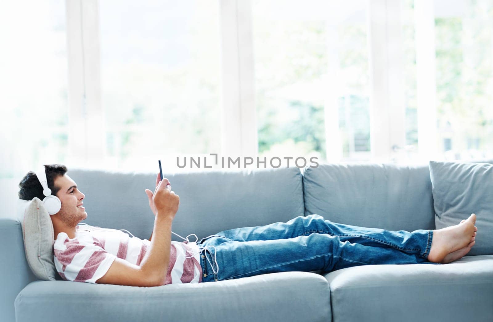 Man, headphones and phone on sofa in house for music, streaming and self care while on study break. Guy, headset and tech on couch in home living room for social media, video and app or radio by YuriArcurs