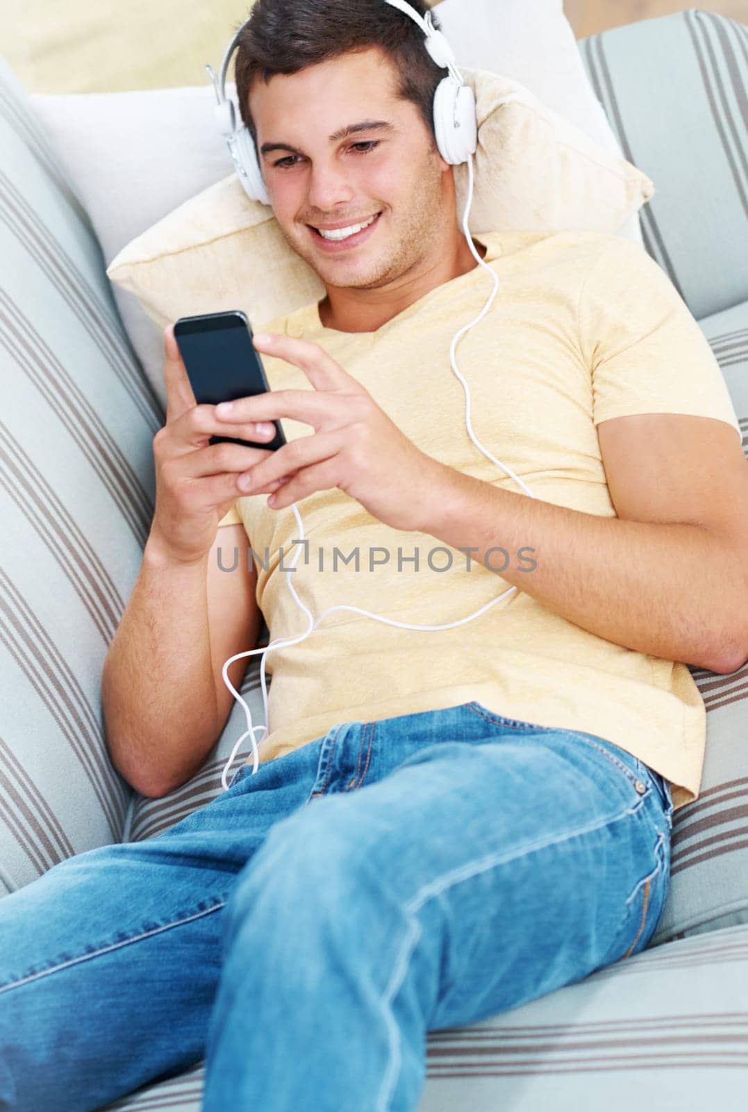 Happy man, headphones and streaming with phone in relax on sofa for music, podcast or audio at home. Young male person with smile on mobile smartphone for online entertainment on couch at house by YuriArcurs