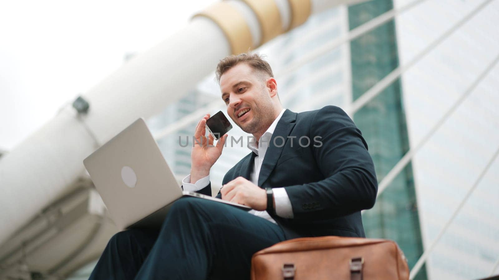 Smart caucasian business man sitting at stairs while using laptop to type to project manager and calling phone to present and plan marketing strategy surrounded by urban city view. Lifestyle. Urbane.
