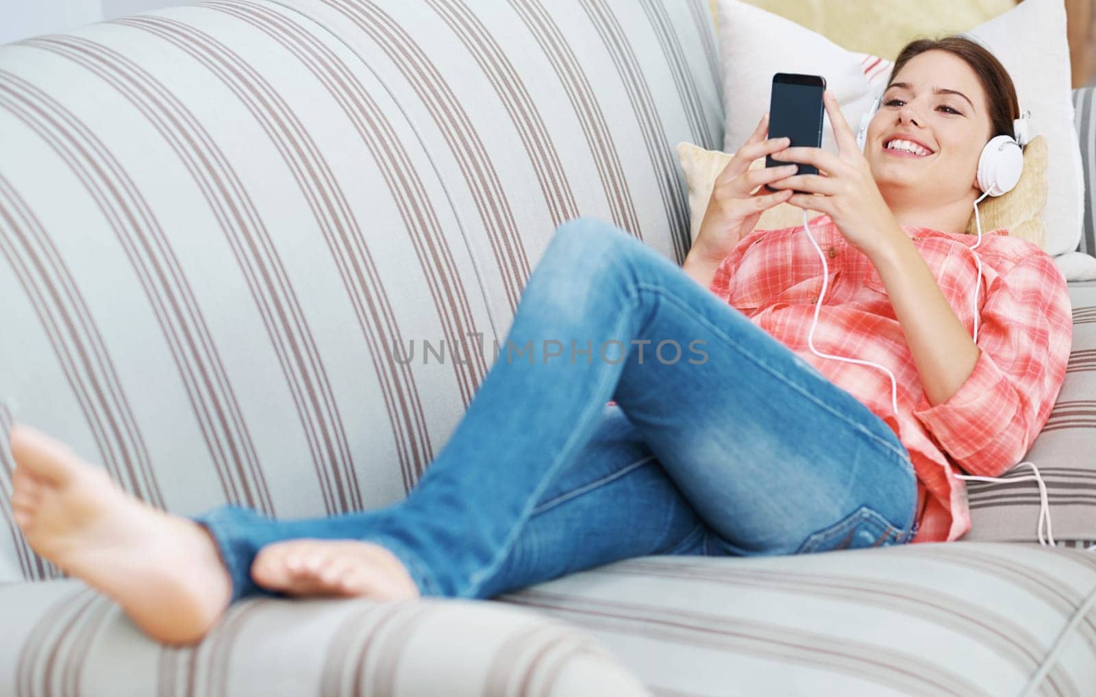 Happy woman, headphones and streaming with phone in relax on sofa for music, podcast or audio at home. Young female person with smile on mobile smartphone for online entertainment on couch at house.