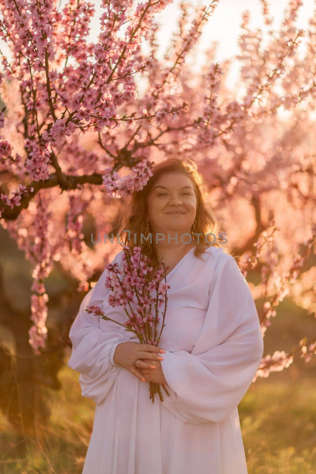 Woman blooming peach orchard. Against the backdrop of a picturesque peach orchard, a woman in a long white dress enjoys a peaceful walk in the park, surrounded by the beauty of nature