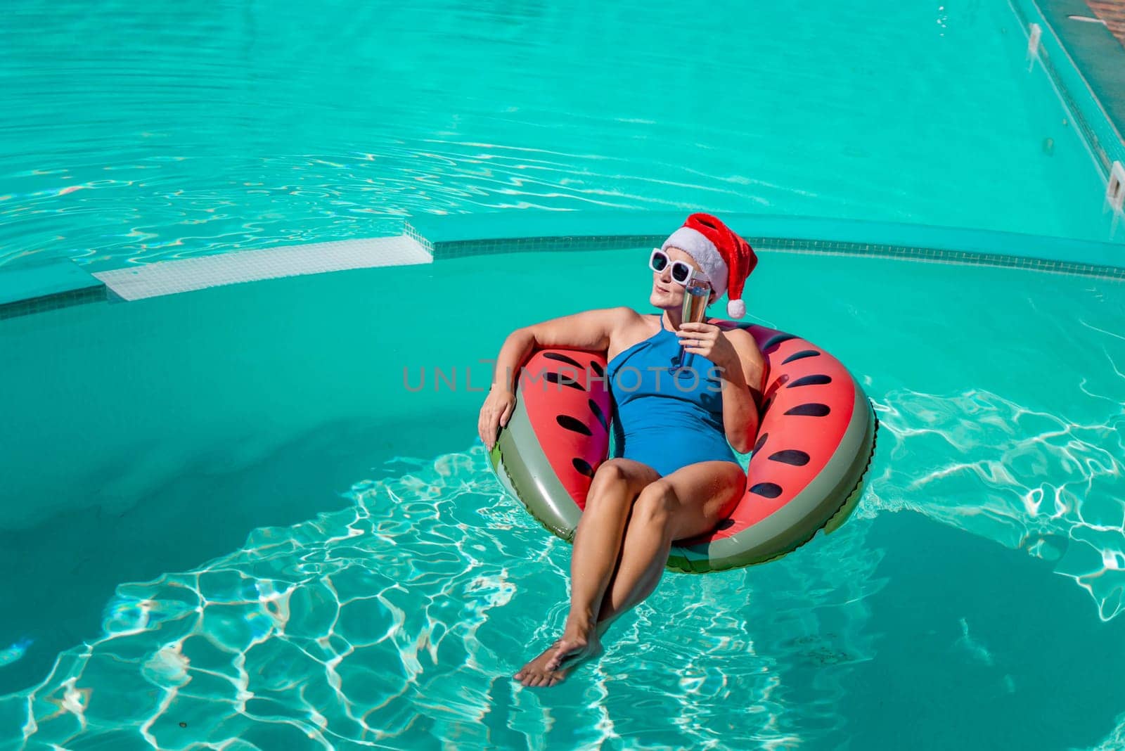 A happy woman in a blue bikini, a red and white Santa hat and sunglasses poses in the pool in an inflatable circle with a watermelon pattern, holding a glass of champagne in her hands. Christmas holidays concept. by Matiunina