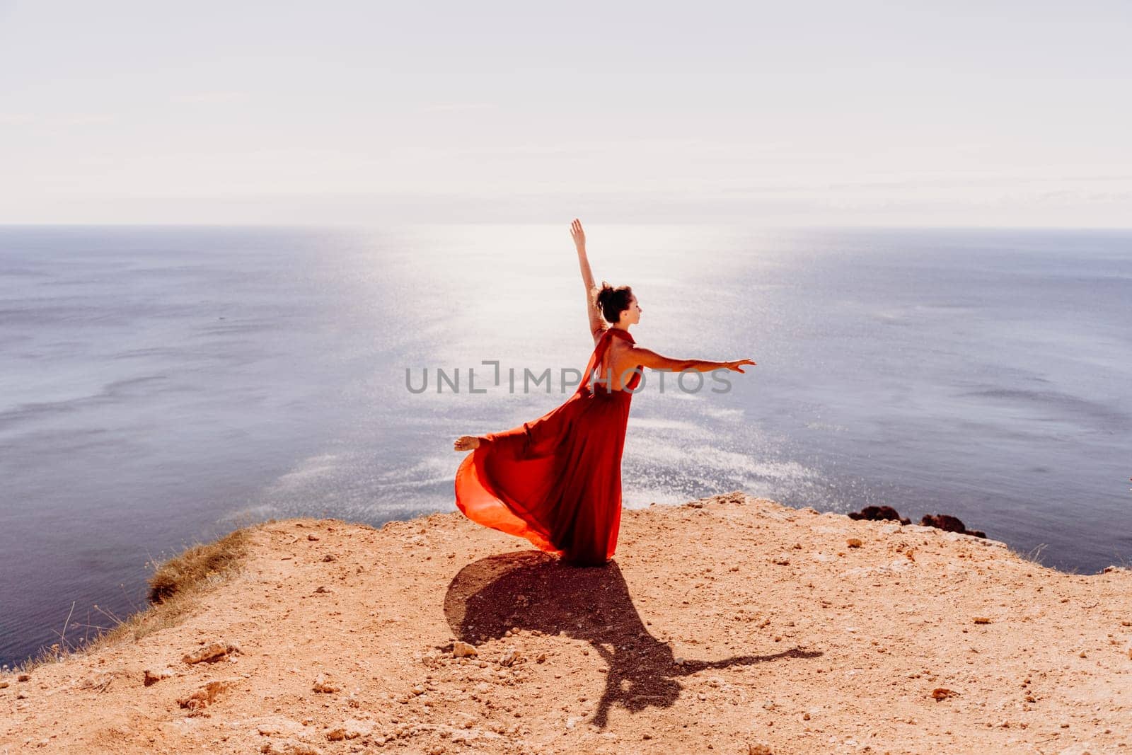 Woman red dress sea. Female dancer posing on a rocky outcrop high above the sea. Girl on the nature on blue sky background. Fashion photo. by Matiunina