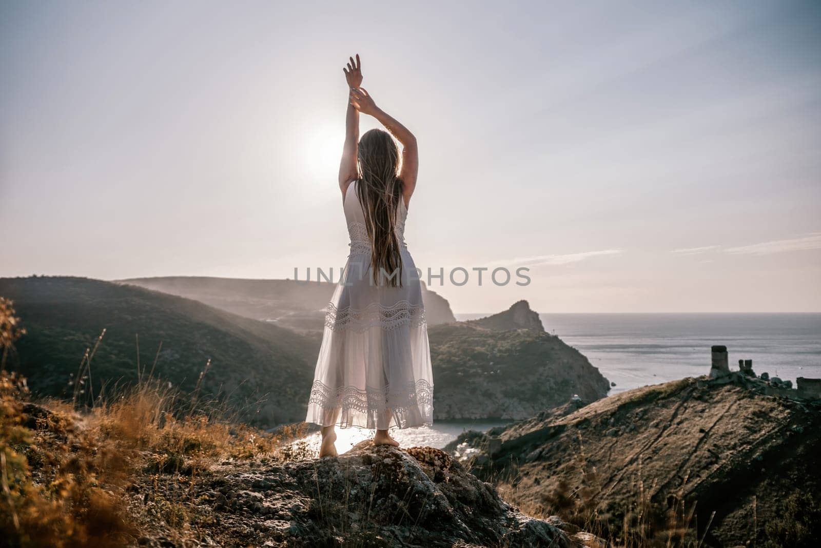 A woman stands on a hill overlooking the ocean. She is wearing a white dress and has her hands on her head. The scene is serene and peaceful, with the woman looking out over the water