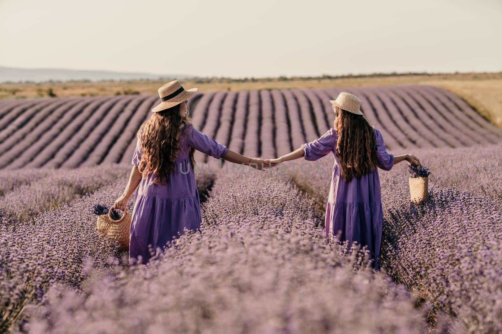 Mom and daughter are running through a lavender field dressed in purple dresses, long hair flowing and wearing hats. The field is full of purple flowers and the sky is clear. by Matiunina