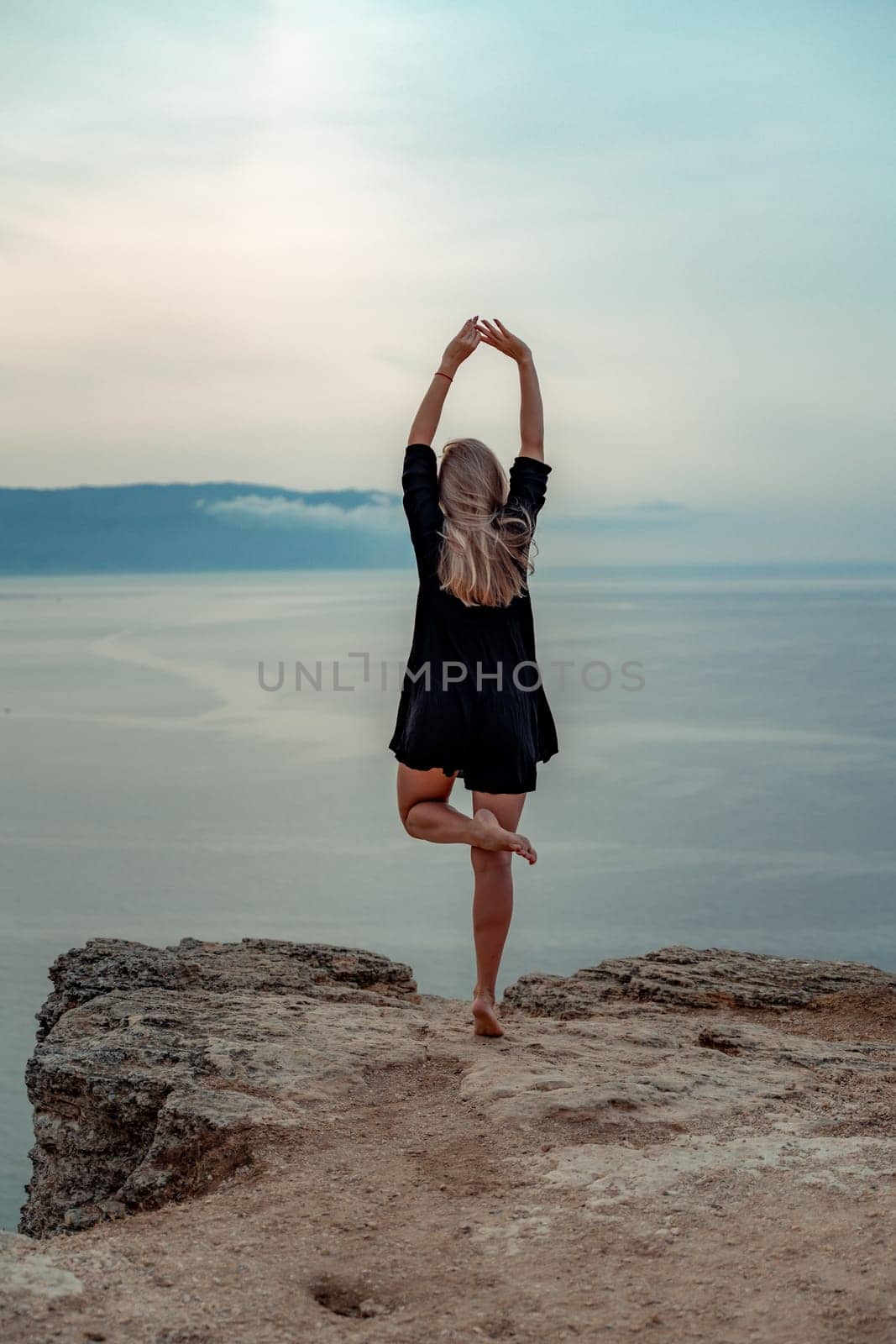 woman stands on a rocky cliff overlooking the ocean. She is wearing a black dress and bare feet. The scene is serene and peaceful, with the woman's presence adding a sense of calm to the image. by Matiunina