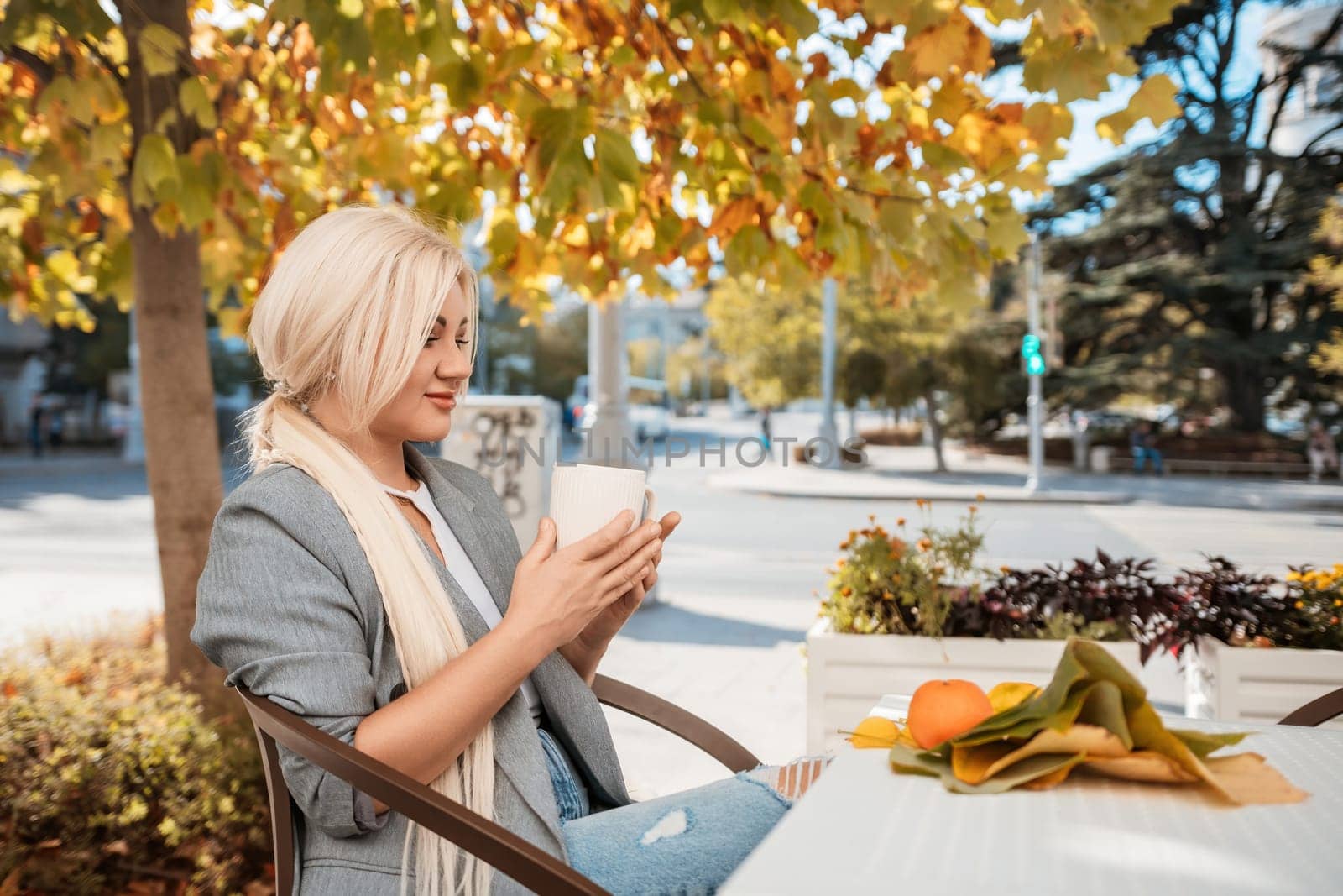 A blonde woman is sitting at a table with a cup of coffee and a plate of fruit. She is smiling and enjoying her time outside. by Matiunina