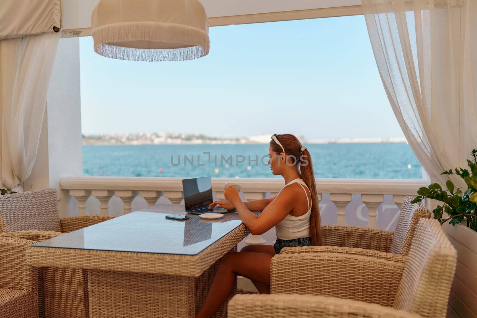 Woman coffee cafe laptop sea. Female freelancer using laptop on coffee break in modern cafe with sea view in the morning by Matiunina