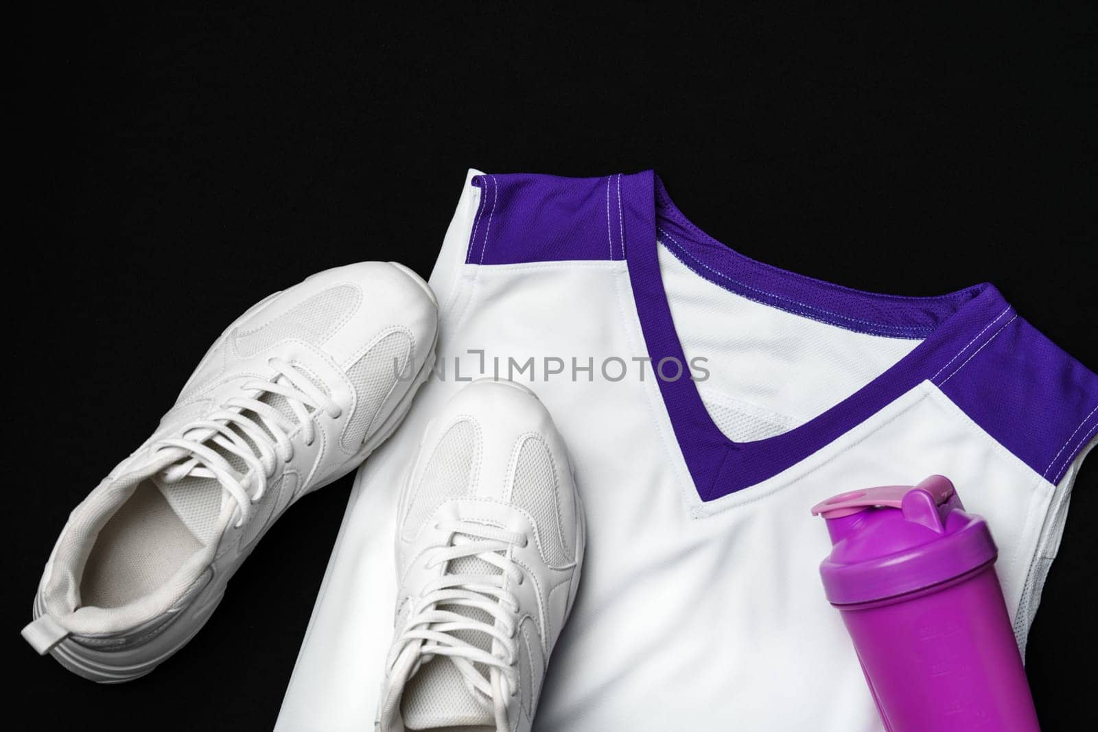 A pair of white sneakers with a purple and white tank top are laid out neatly next to a purple shaker bottle, all arranged against a stark black backdrop, suggesting a preparedness for a fitness or athletic endeavor.