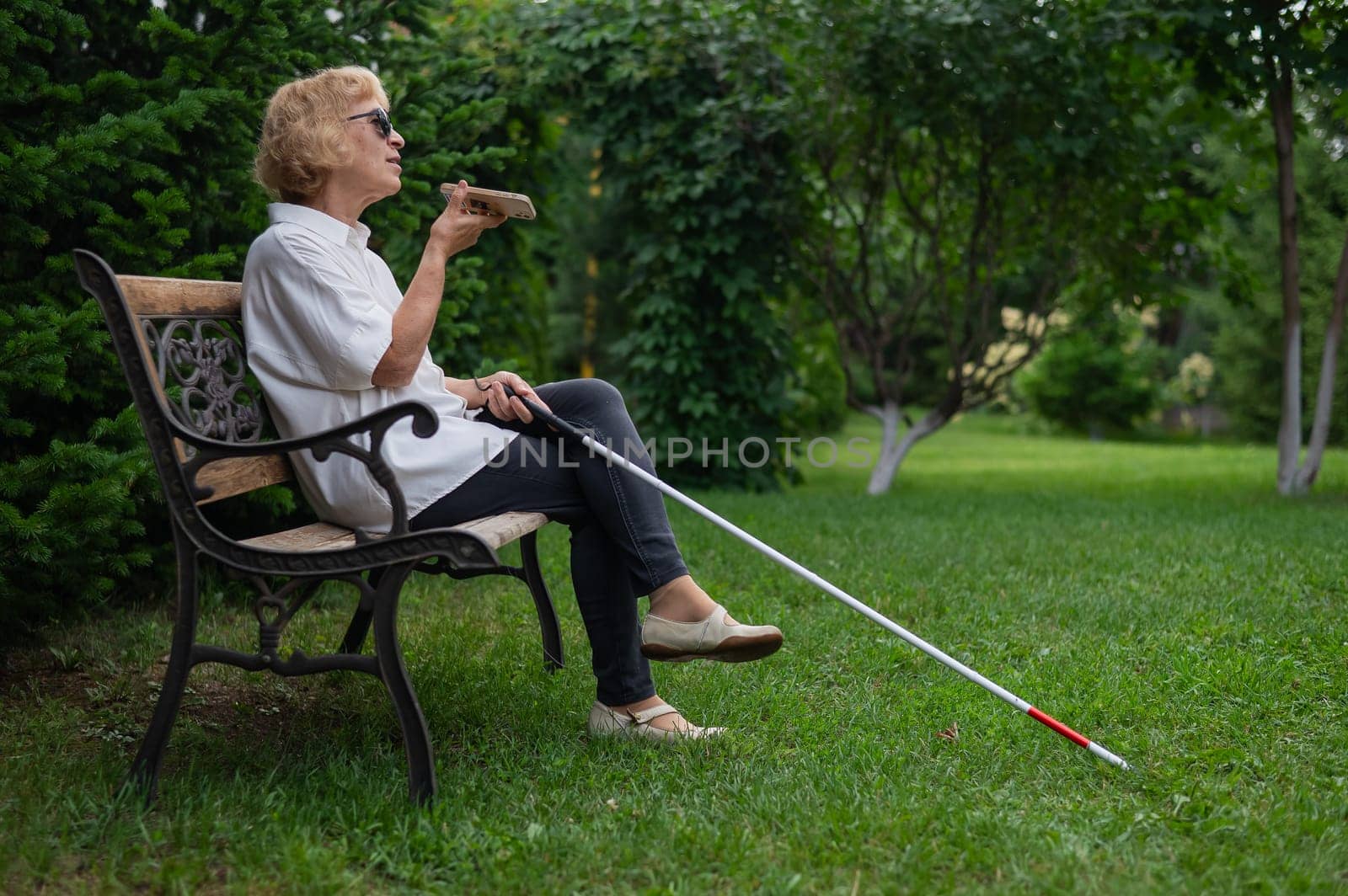 Elderly blind woman talking on a smartphone while sitting on a bench in the park. by mrwed54