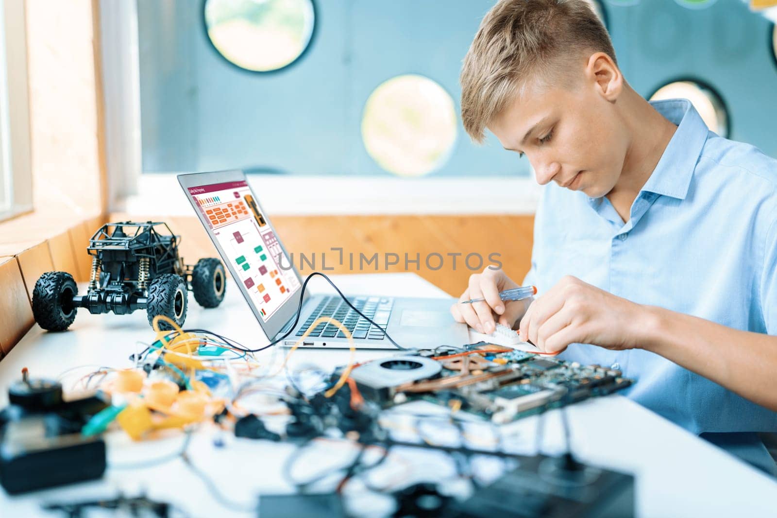 Schoolboy in blue shirt connect parts of robotics vehicle with motherboard carefully in STEM class. On table put laptop, controller, electric wire, battery charger, and robotic vehicle. Edification.