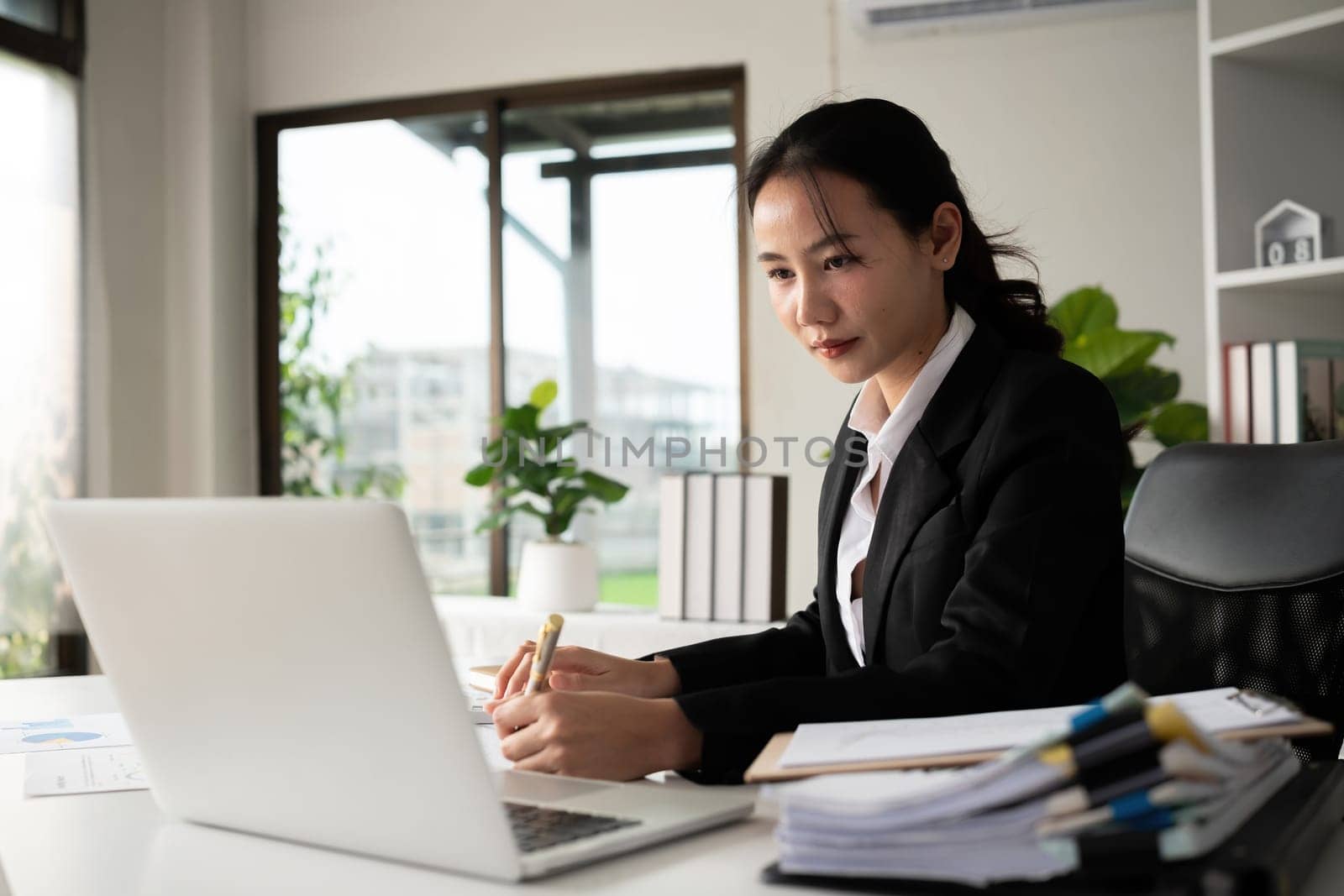 Focused Asian female accountant doing paperwork in office with plan documents on desk by wichayada