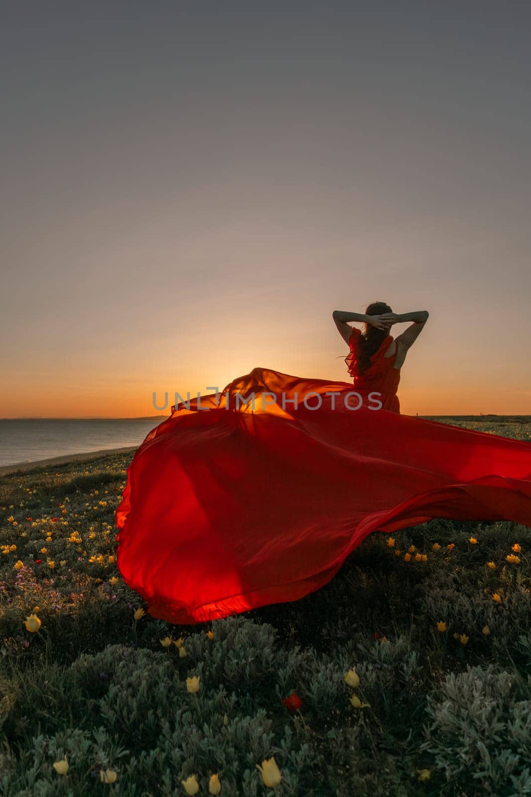 A woman in a red dress is standing in a field with the sun setting behind her. She is reaching up with her arms outstretched, as if she is trying to catch the sun. The scene is serene and peaceful. by Matiunina