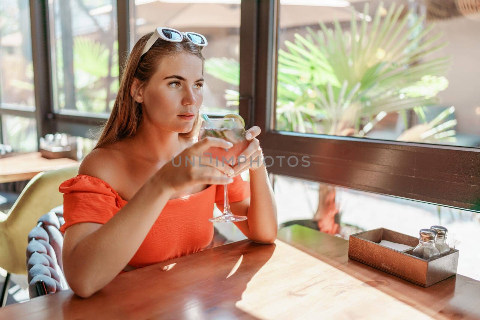 woman in a cafe seated, holding beverage. Dressed in an orange dress with her hair down. Bright interior, large windows allow natural light. Beverage consumption for refreshment. by Matiunina