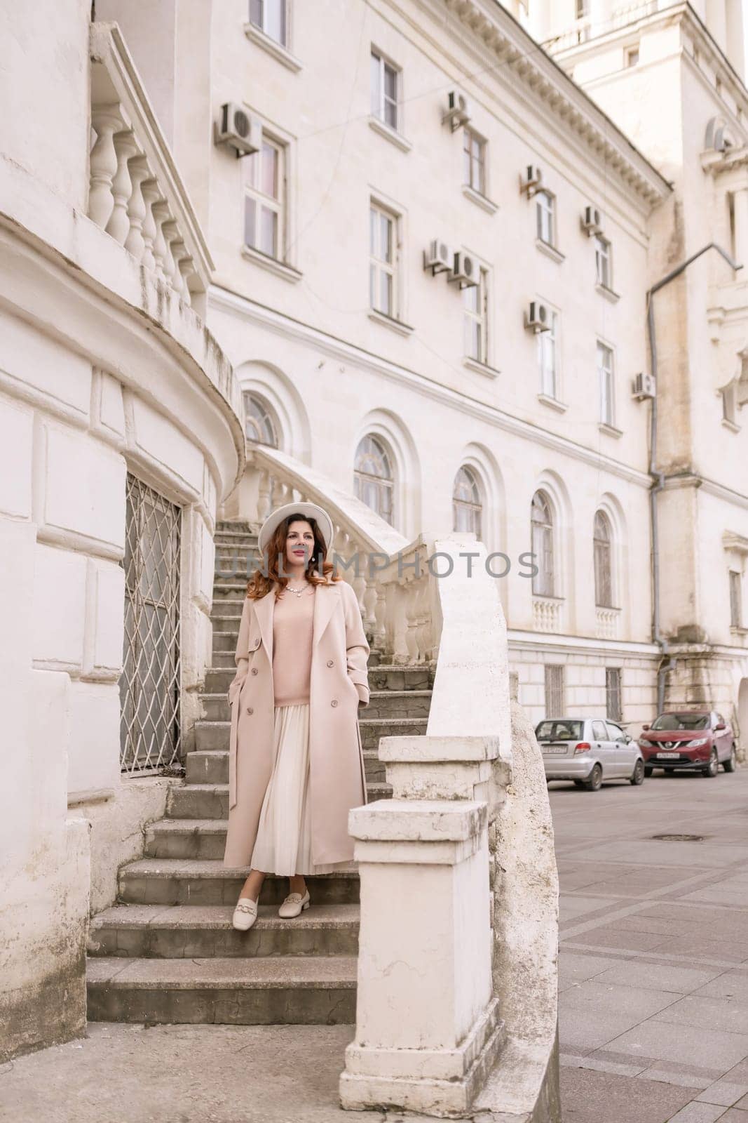 woman in elegant coat and hat against an intricate architectural backdrop, harmoniously blending modern fashion with historical allure. The soft daylight adds to its timeless appeal. by Matiunina