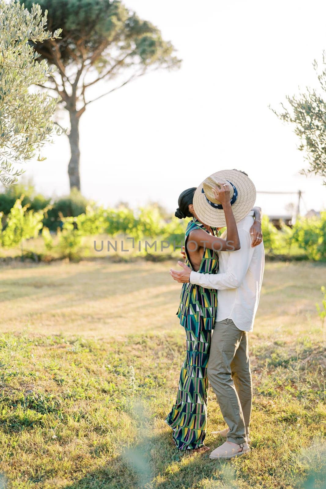 Man kisses woman while standing in a green garden and hiding behind a straw hat by Nadtochiy