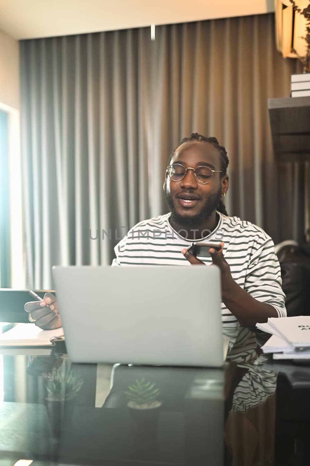 Smiling African American man in eyeglasses using laptop and talking on mobile phone at home.