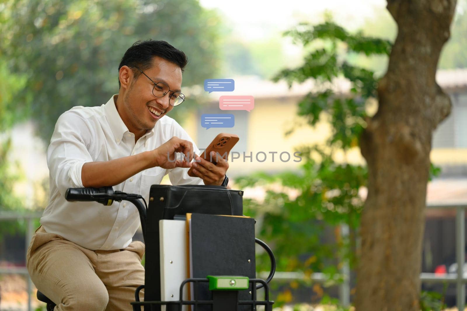 Pleased young businessman using mobile phone while sitting on a bicycle outdoor by prathanchorruangsak