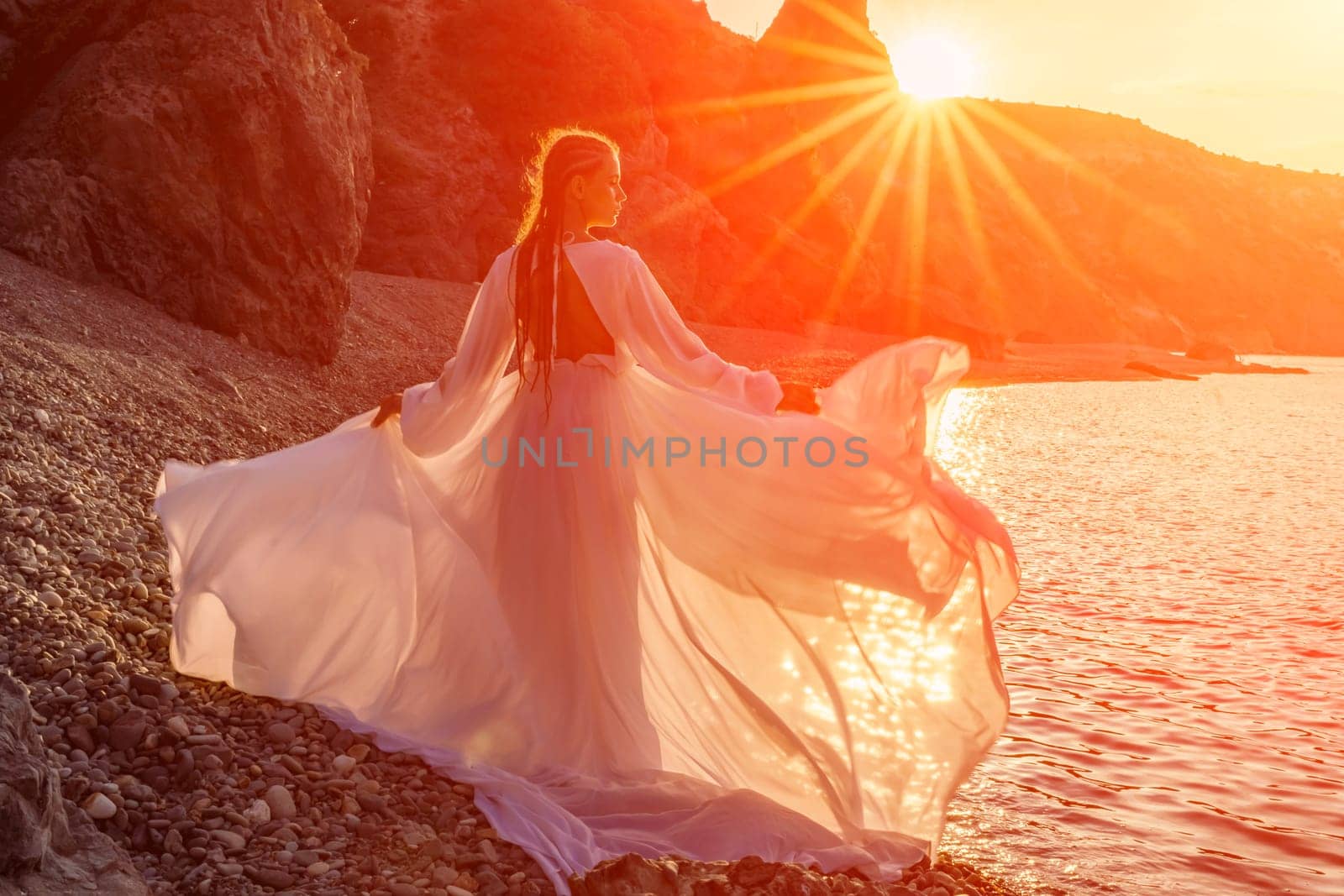Mysterious woman silhouette long hair walks on the beach ocean water, sea nymph wind listens to the wave. Throws up a long white dress, a divine sunset. Artistic photo from the back without a face by Matiunina