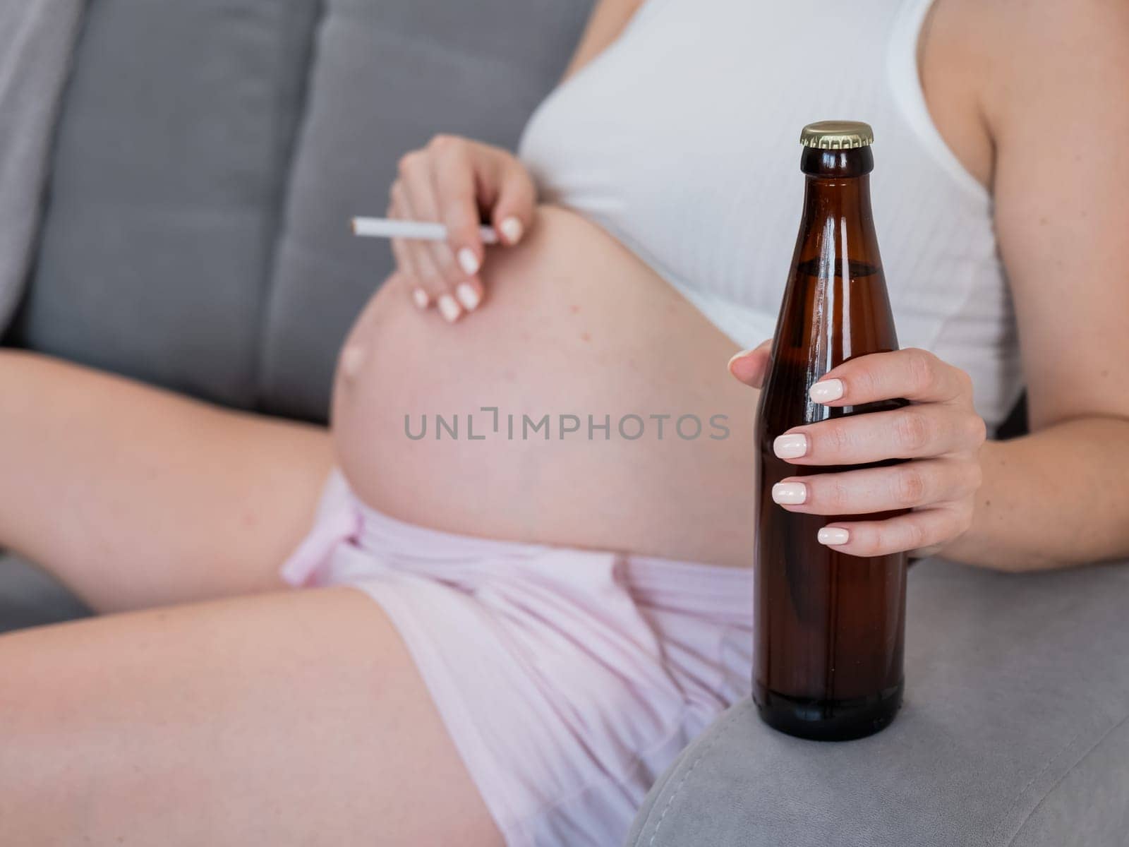 Faceless pregnant woman drinks beer while sitting on the sofa