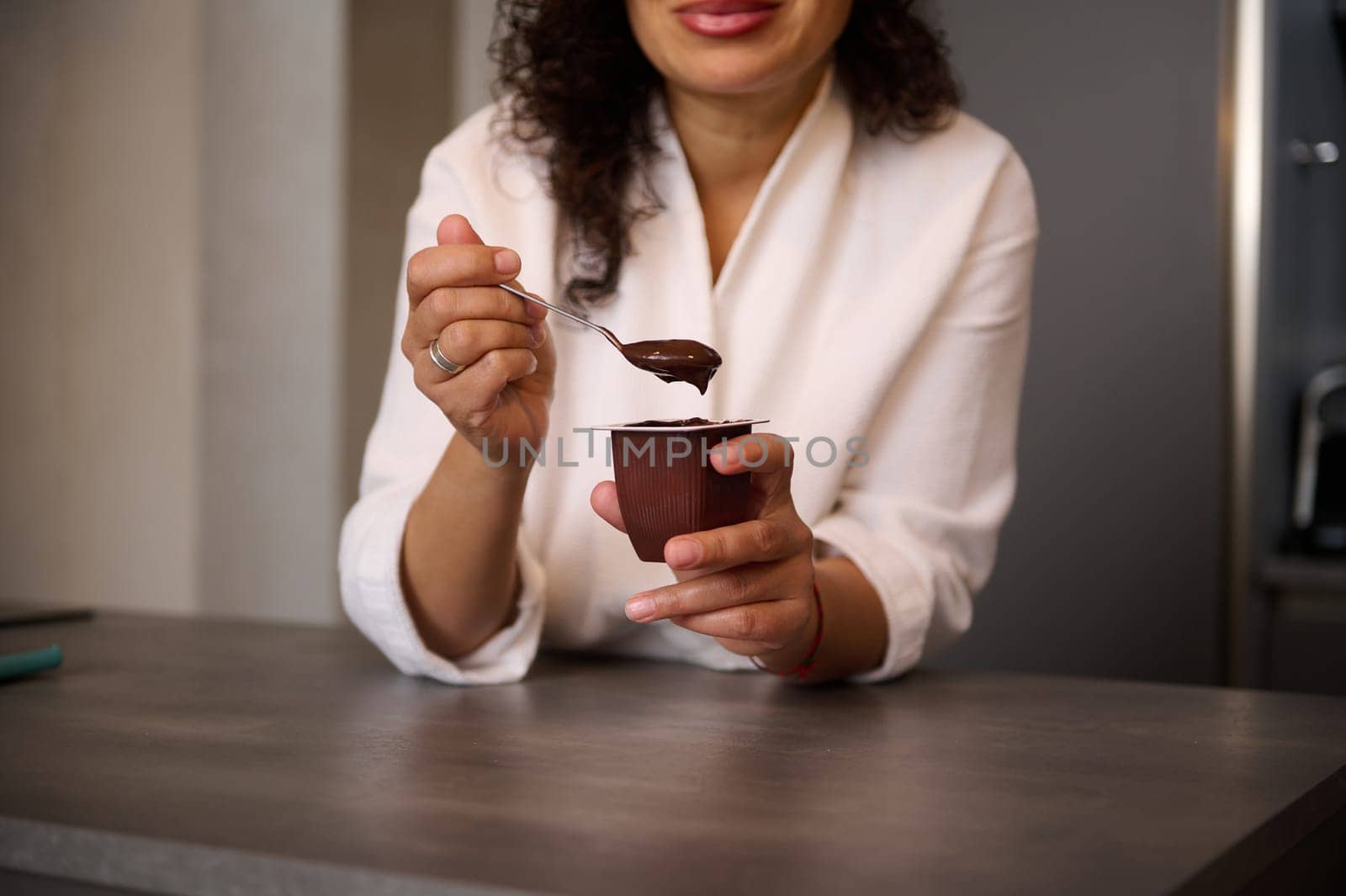 Selective focus on woman's hands, holding a teaspoon and a container with wholesome chocolate vegetarian yoghourt for breakfast. Healthy food and diet concept