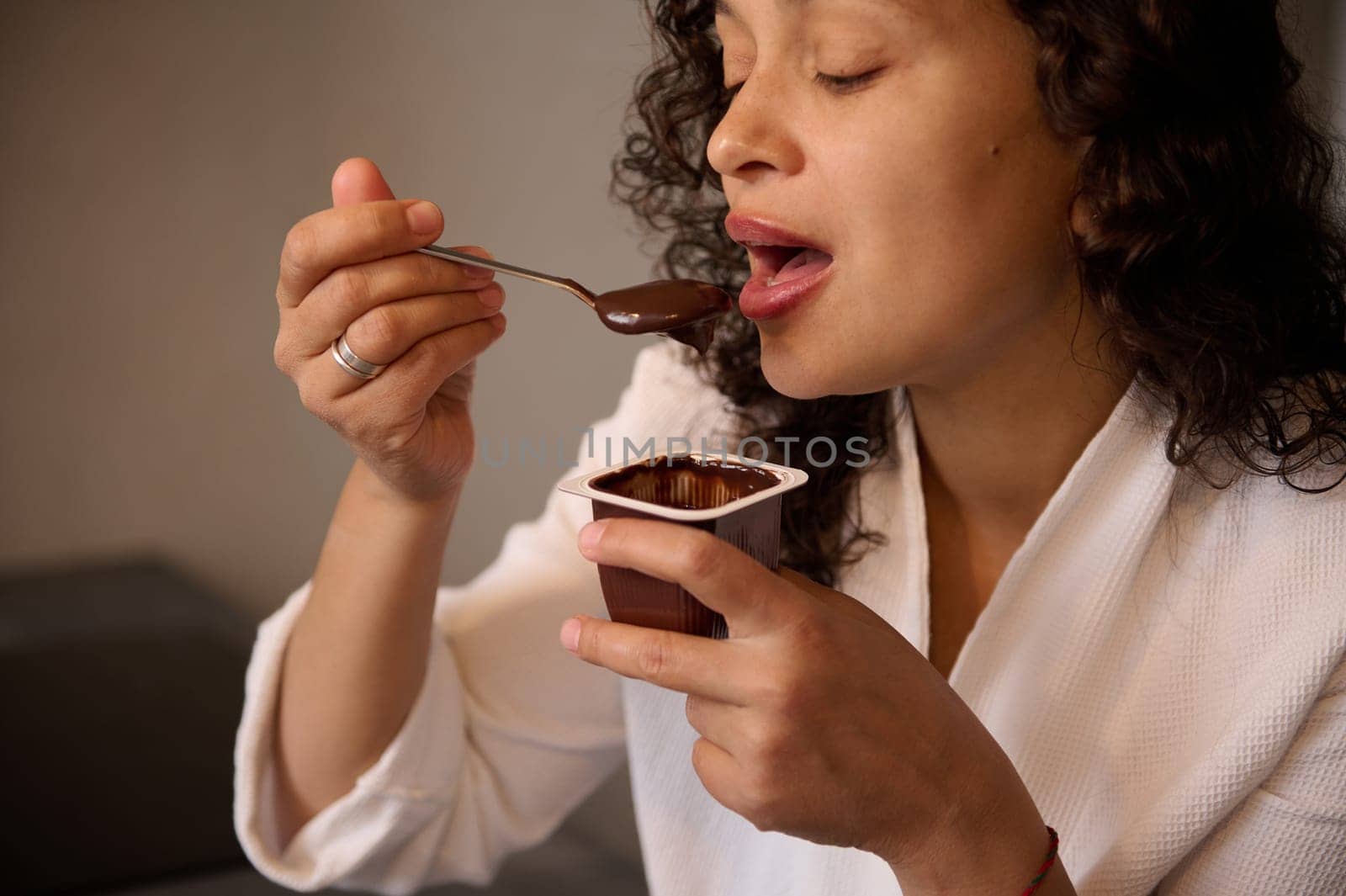 Authentic portrait of curly haired brunette woman enjoying the taste of chocolate vegan yogurt. Multi ethnic pretty female snacking with tasty yoghurt at home. Food and drink consumerism. Diet concept